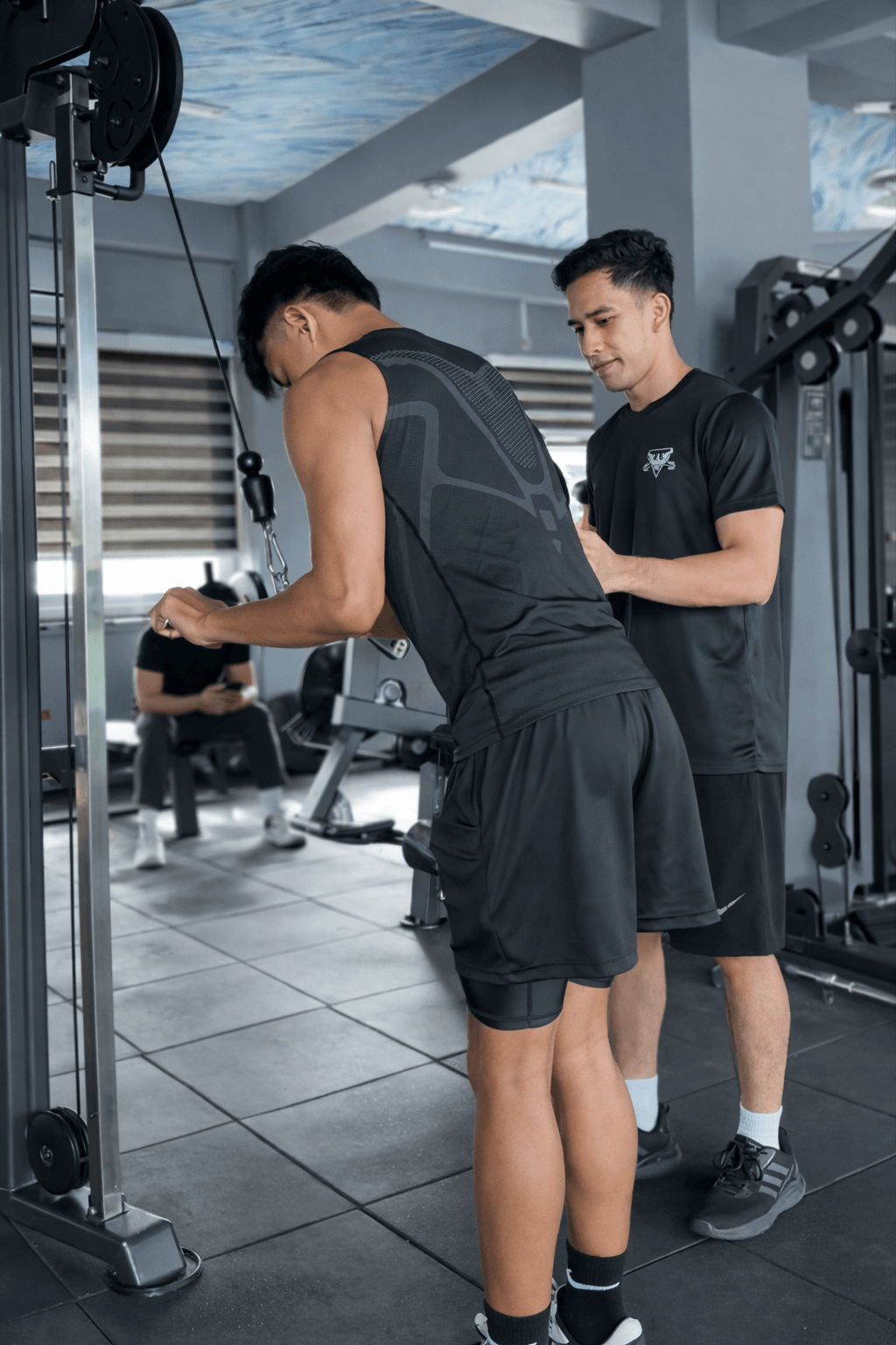 A Power Flex coach checks a member's form on the cable pushdown, one hand on the member's back.
